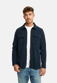 Mørkeblå kordfløjlsjakke med lynlåsfortil, to brystlommer og lange ærmer, båret over en hvid T-shirt og lyseblå jeans.