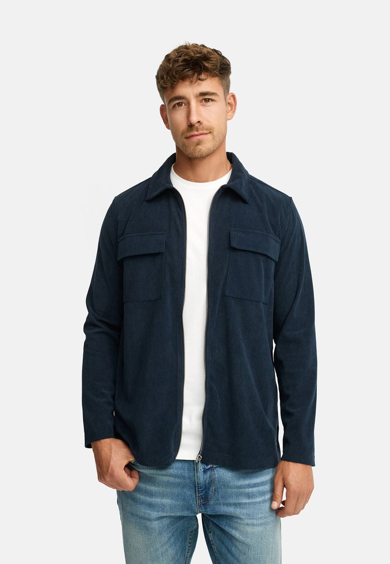 Mørkeblå kordfløjlsjakke med lynlåsfortil, to brystlommer og lange ærmer, båret over en hvid T-shirt og lyseblå jeans.