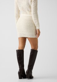 Bottes hautes en cuir marron à talon carré, avec un détail de fermeture éclair dorée et une finition texturée, complétées par une robe en maille crème.