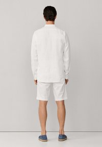 Hombre de pie de espaldas, vistiendo una camisa blanca de manga larga, pantalones cortos blancos y zapatos azules sin cordones, frente a un fondo liso.