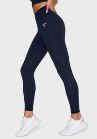 Marinblå leggings gjorda av stretchigt material med hög midja, med en sidologga och sömdetaljer. Bärs med vita träningsskor.