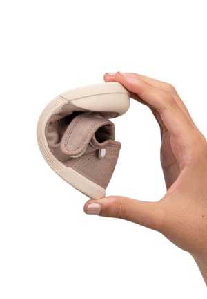 Zapato de tela beige flexible sostenido en una mano, con punta redondeada, textura suave y suela de goma blanca contrastante.