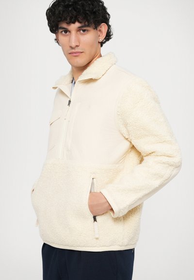 Homem vestindo uma jaqueta de fleece creme com bolsos com fecho, fecho frontal até a metade e tecido texturizado nas mangas e no corpo.