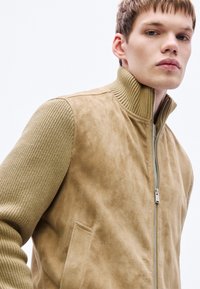 Beežikas sinepikollase varrukate ja kõrge kraega sametjakk. Esiosas lukuga ja küljetaskud, sile pind. Muster puudub.