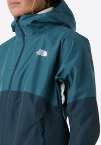 Blue and dark teal waterproof jacket with a hood, zippered pockets, and a soft inner lining. Features a small logo on the chest.