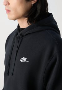 Musta Nike huppari, jossa on etutasku, nauhakuulokkeet ja brodeerattu valkoinen logo vasemmassa rinnassa. Pehmeä, teksturoitu kangas.