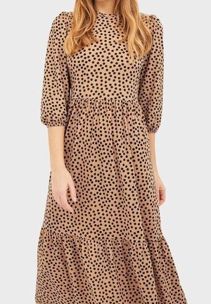 Vrouw die een zandkleurige midi-jurk draagt met zwarte polka dots, driekwart pofmouwen en een geplooide taille, tegen een effen lichte achtergrond.