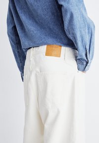 Fehér pamut nadrág hátsó zsebekkel, világosbarna bőrcímkével, amelyen az "Jack & Jones" felirat áll. Kék chambray inggel párosítva.