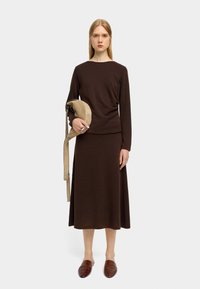 Brązowy sweter o gładkiej teksturze i dopasowana spódnica, długie rękawy, okrągły dekolt. Modelka trzyma beżową torebkę na ramię i nosi brązowe klapki.