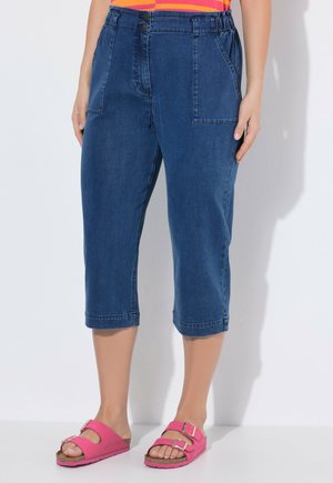 Blå denimbukser i midtlængde med forlommer båret af en person i lyserøde sandaler med dobbeltspænde mod en ensfarvet baggrund.