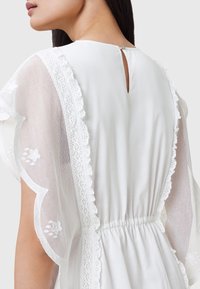 Abito bianco con maniche in tessuto trasparente rifinite in pizzo ed ricamo, chiusura a fessura sul retro e vita elastica per un look aderente.