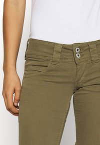 Olivově zelené, elastické bavlněné kalhoty s úzkým střihem a zapínáním na dva knoflíky. Mají přední kapsy a viditelné švy.