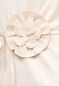 Λεπτομέρεια λουλουδιού από δέρμα χρώματος κρέμα, προσκολλημένη σε ζώνη από υφασμάτινο υλικό του ίδιου χρώματος, πάνω σε ρούχο ανοιχτού μπεζ χρώματος.