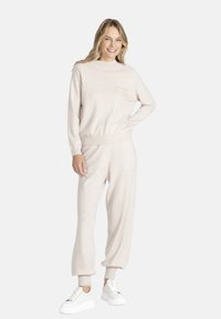 Jasno beżowy sweter i dopasowane dresy wykonane z miękkiej dzianiny. Sweter ma okrągły dekolt i kieszonkę na klatce piersiowej; dresy mają ściągacze przy kostkach.