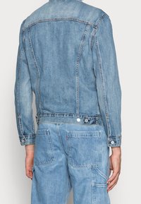 Jasnoniebieska kurtka dżinsowa o lekko wyblakłej teksturze, widocznymi szwami, detalami z guzikami i dopasowanym kroju, pokazana z tyłu.