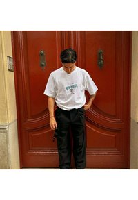 Camiseta gráfica blanca con diseño de dibujos animados, combinada con pantalones plisados negros. El fondo presenta una puerta de madera roja con herrajes decorativos.