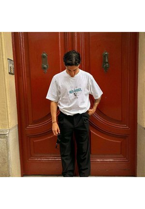 Camiseta gráfica blanca con diseño de dibujos animados, combinada con pantalones plisados negros. El fondo presenta una puerta de madera roja con herrajes decorativos.