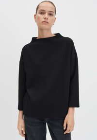 Frau in einem lässigen, schwarzen, strukturierten Langarmoberteil und dunklen Jeans, die vor einem schlichten weißen Hintergrund steht und nach vorne schaut.