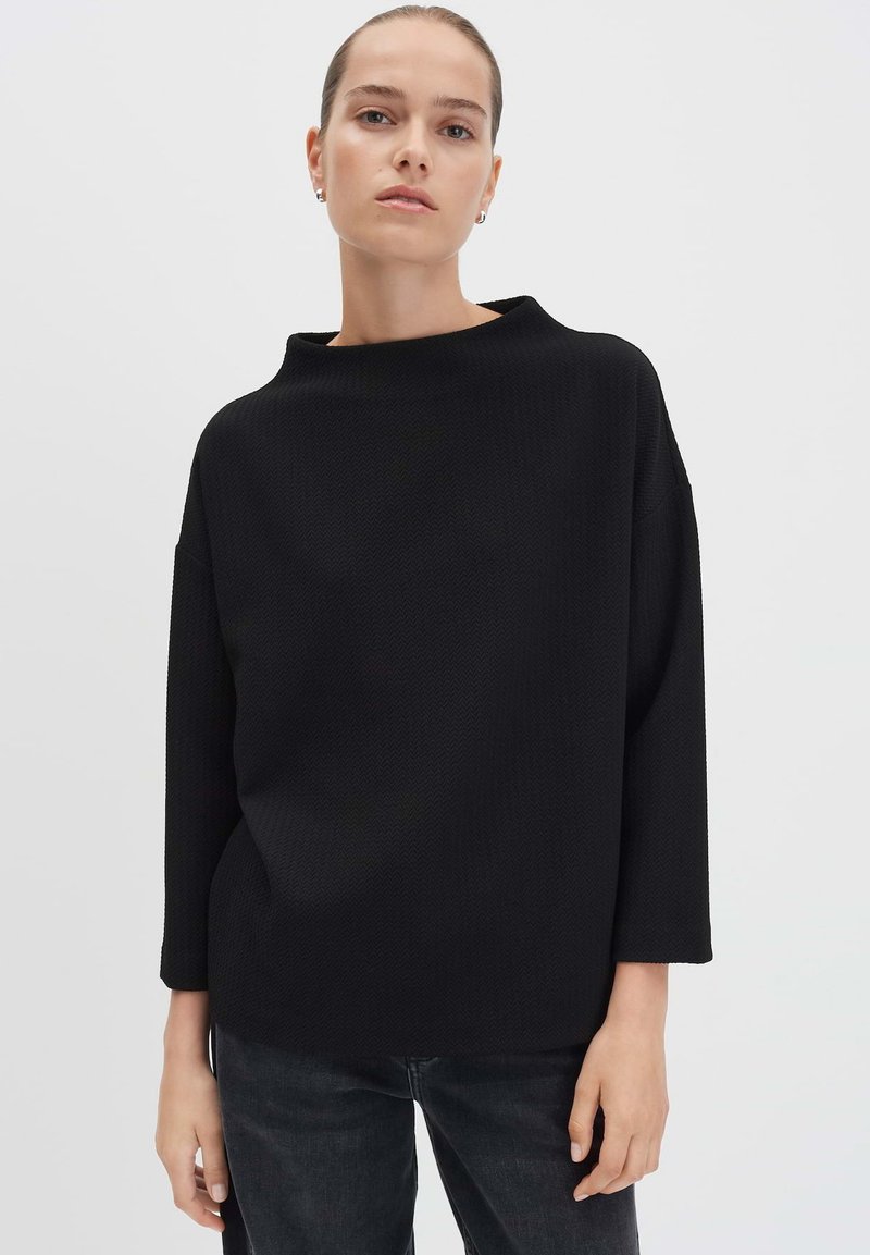 Frau in einem lässigen, schwarzen, strukturierten Langarmoberteil und dunklen Jeans, die vor einem schlichten weißen Hintergrund steht und nach vorne schaut.