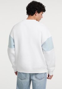 Felpa bianca con pannelli d'accento azzurri, vestibilità rilassata, texture morbida, scollo rotondo e orlo a coste. Indossata con jeans azzurri.