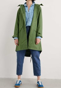 Grön, lätt regnjacka med huva, tryckknappar och två framfickor, lagd över en ljusblå skjorta och blå denimbyxor.