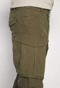 Vue latérale d'un pantalon cargo vert olive avec poche à rabat et fermeture éclair visible au niveau de la hanche.