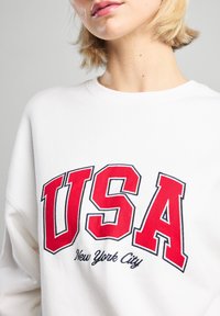 Felpa bianca con grande scritta rossa "USA" e "New York City" in un carattere più piccolo, con collo girocollo e vestibilità comoda.
