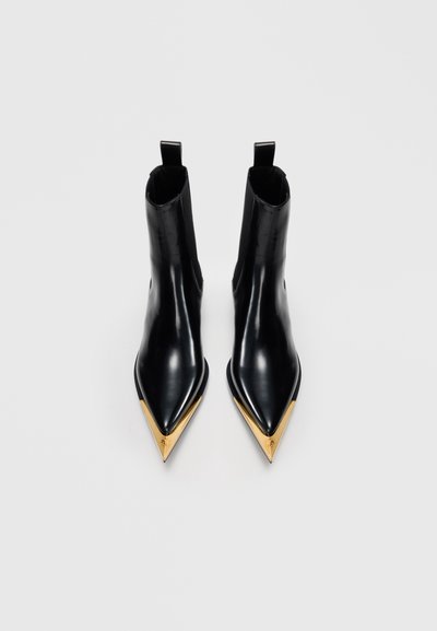 Svart ankelboots med spetsig tå och glänsande guldmetallkapslar på tårna mot en enkel ljus bakgrund.
