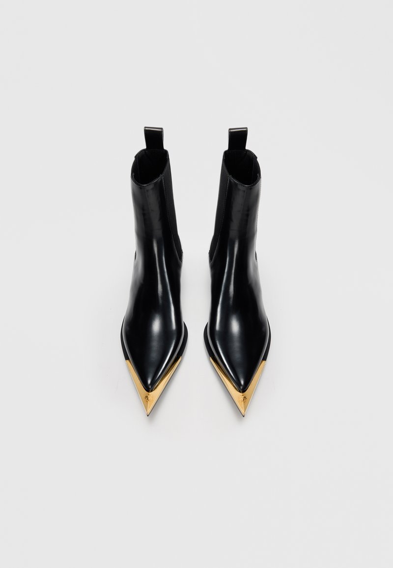Stivaletti neri con punta a punta e tacco corto, dotati di eleganti coperture in metallo dorato sulla punta, su uno sfondo chiaro e uniforme.