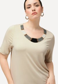 Beige Kurzarmoberteil mit einem dekorativen Ausschnitt, der mit Perlen in Schwarz, Grau, Weiß und goldenen Akzenten verziert ist, aus einem glatten Material gefertigt.