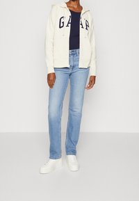 Haut de survêtement zippé beige clair avec le logo "GAP", associé à un jean bleu clair ajusté et des baskets blanches. Tissu lisse et design décontracté.