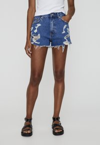 Shorts de mezclilla en azul claro con detalles desgastados, dobladillo desflecado, diseño de cintura alta y dos bolsillos frontales. Combinados con sandalias negras.