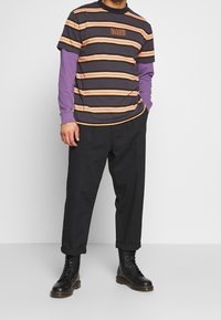 Stribet t-shirt i mørkegrå, orange og gul over en lilla langærmet skjorte, parret med sorte tapered bukser og sorte snørebåndsstøvler.