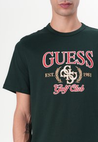 Camiseta de algodón verde oscuro con un logotipo bordado en rojo y oro y el texto "GUESS EST. 1981 Golf Club" con elementos decorativos.
