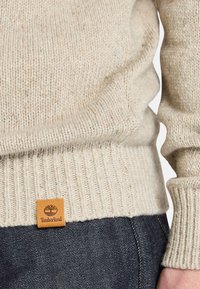 Beżowy sweter z dzianiny z prążkowanymi mankietami i dolnym brzegiem; ma brązową skórzaną metkę Timberland na dolnym lewym boku.