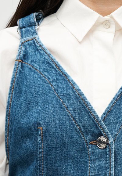 Λευκό πουκάμισο με γιακά κάτω από μπλε φόρεμα σαλοπέτα από τζιν με μεταλλικά κουμπιά και λεπτομέρειες καφέ ραφής.