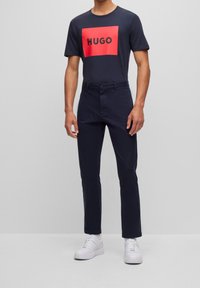 Navy T-Shirt mit rotem rechteckigem Logo mit der Aufschrift "HUGO" in der Mitte, kombiniert mit navyfarbigen Hosen und weißen Sneakers. Minimalistisches Design.