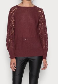 Bordeauxroter Pullover mit gestricktem Rumpf und Spitzenärmel. Ausgestattet mit einem runden Halsausschnitt und gerippten Bündchen. Kombiniert mit schwarzen, figurbetonten Hosen.