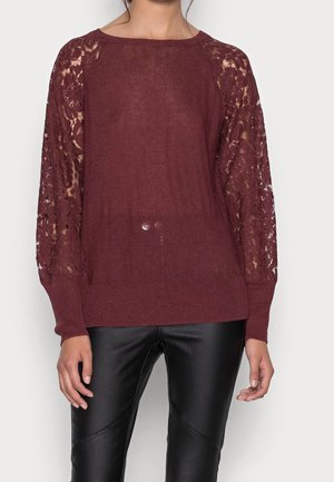 Pull bordeaux avec un corps tricoté et des manches en dentelle. Il présente un col rond et des poignets côtelés. Associé à un pantalon noir ajusté.
