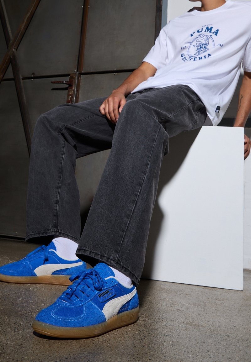Personne portant des baskets Puma bleu vif avec des chaussettes blanches, un pantalon large noir et un t-shirt blanc Puma, assise sur un bloc blanc.