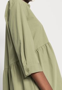 Robe en coton vert clair avec un col rond, des manches trois-quarts, un corsage froncé et des détails de poignet à boutons. Texture lisse.