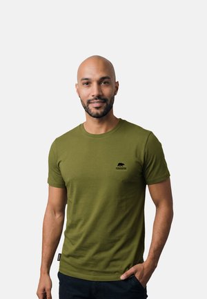 Mann mit kurzem Bart, der ein olivgrünes Forsberg-T-Shirt und dunkle Hosen trägt, steht mit einer Hand in der Tasche vor einfarbigem Hintergrund.