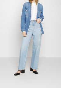 Chaqueta de mezclilla azul clara con bordes deshilachados, camisa blanca de botones, jeans azul claro de cintura alta y zapatos negros de tacón cuadrado.