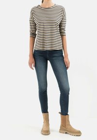 Woman wearing beige and black striped long-sleeve top, dark blue skinny jeans, and beige suede ankle boots, standing on white background.