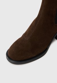 Bota de tobillo de ante marrón con punta redondeada, un bajo tacón de goma negra y laterales elásticos para facilitar su puesta. Textura suave.
