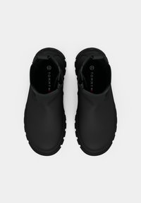 Des chaussures noires à enfiler fabriquées en matériau lisse et imperméable, avec une semelle en caoutchouc texturée et un marquage minimal à l'intérieur.