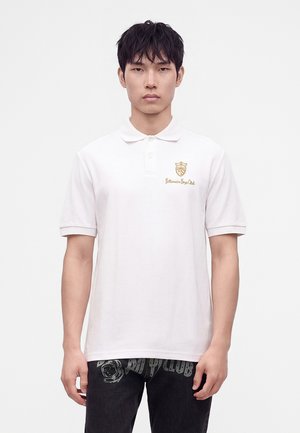 Jeune homme portant un polo blanc avec un emblème doré et un texte sur la poitrine, associé à un pantalon noir avec des motifs imprimés.