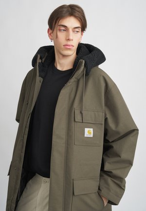 Olivengrüner Jacke mit einer schwarzen, fleecegefütterten Kapuze, einem vorderen Reißverschluss und einer Brusttasche mit einem Logo-Patch. Über einem schwarzen Shirt getragen.