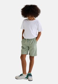 T-shirt en coton blanc associé à un short vert menthe avec une taille élastique. Baskets blanches avec des sangles bleues et vertes.