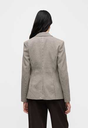 Kvinde med langt mørkt hår iført en tætsiddende ternet blazer og mørke bukser, stående med ryggen til mod en ensfarvet lys baggrund.